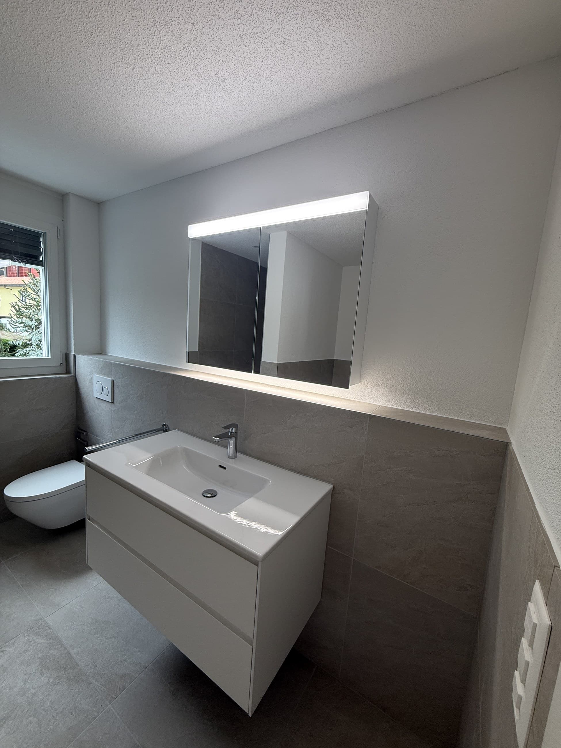 Graues Badezimmer mit Spiegelschrank, weissem Waschtisch und Tageslicht vom Fenster