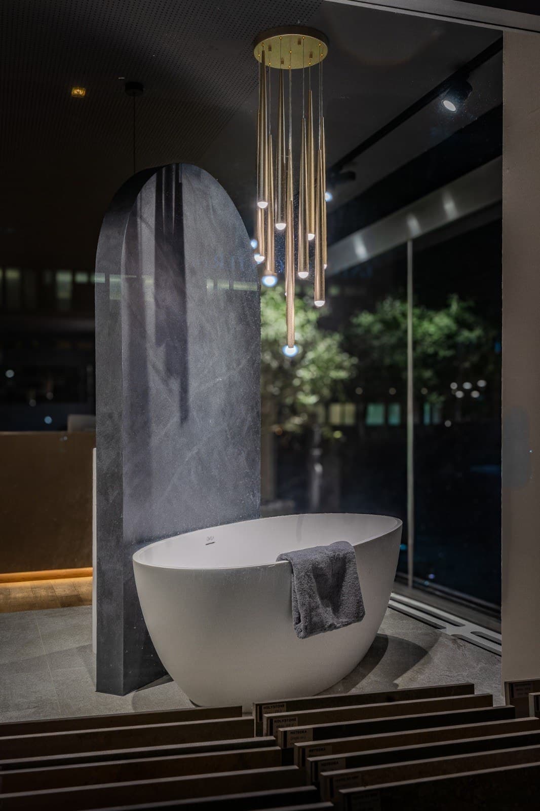 Freistehende Badewanne vor einer Marmorstele mit Goldpendelleuchten im OVEA Showroom Baden